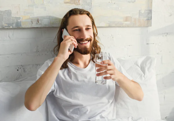 Fröhlicher bärtiger Mann mit langen Haaren, der am Smartphone spricht und morgens zu Hause im Bett Wasser trinkt — Stockfoto