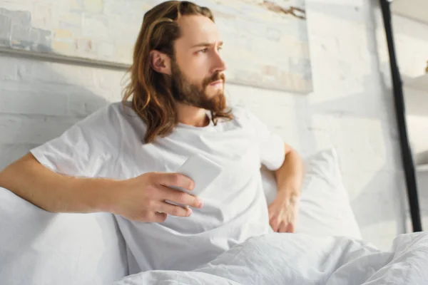 Selective focus of confident man with long hair holding smartphone and looking away in bedroom at home — Stock Photo