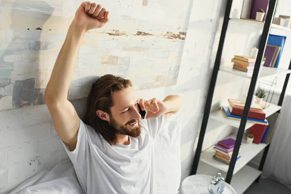 Vue grand angle de l'homme barbu avec les cheveux longs faire étirement et parler sur le lit smartphone pendant le matin à la maison — Photo de stock