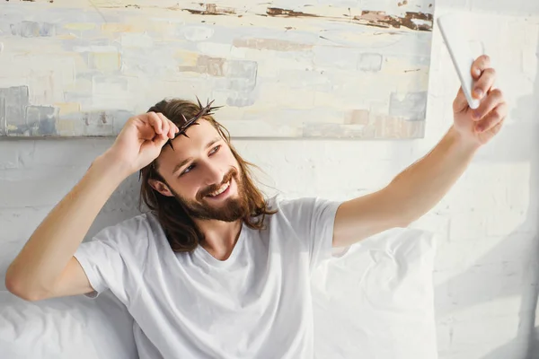 Feliz Jesús en corona de espinas tomando selfie en la cama durante la mañana en casa - foto de stock