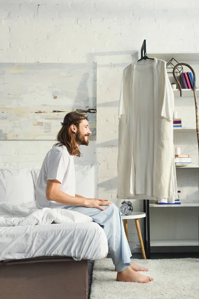 Vista laterale di Gesù felice seduto sul letto durante il mattino in camera da letto a casa — Foto stock