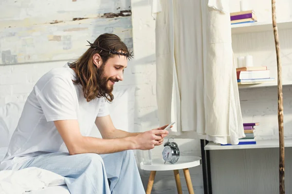 Cheerful Jesus in crown of thorns using smartphone in bedroom at home — Stock Photo