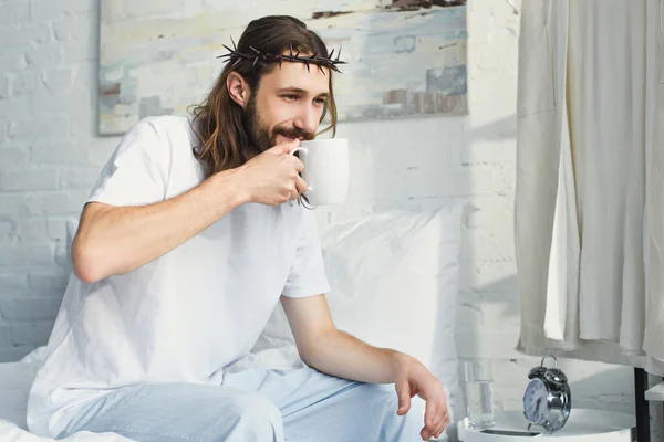 Selective focus of Jesus in crown of thorns drinking coffee in bedroom during morning time at home — Stock Photo