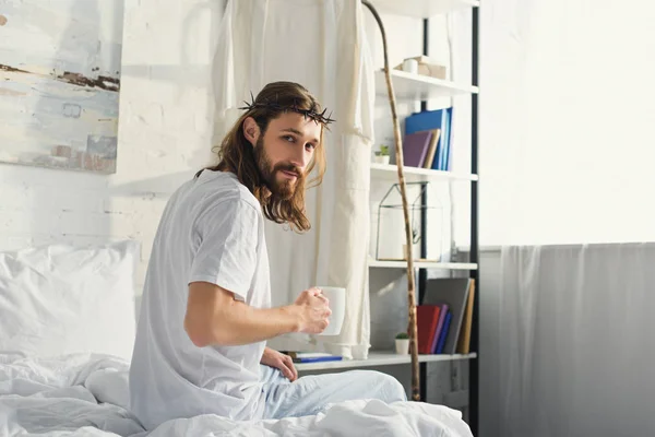 Selective focus of Jesus in crown of thorns looking at camera and holding coffee cup in bedroom during morning time at home — Stock Photo