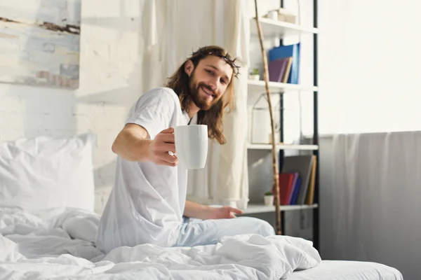 Selective focus of Jesus in crown of thorns showing coffee cup in bedroom at home — Stock Photo