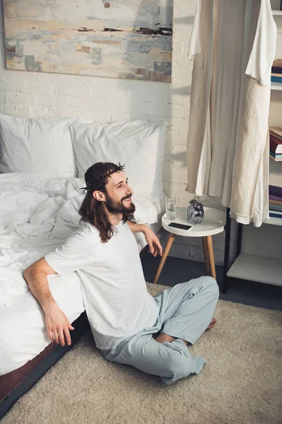 Висока кут зору посміхаючись Ісуса сидить на підлогу біля ліжка під час ранкової час в спальні в домашніх умовах — стокове фото