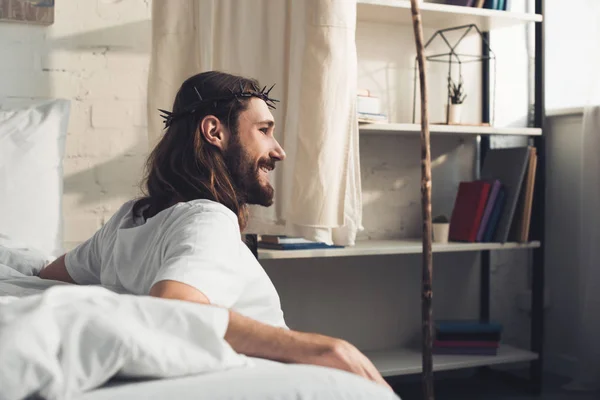 Вид сбоку улыбающегося Иисуса, сидящего на полу возле кровати в спальне дома в утреннее время — стоковое фото