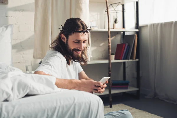 Счастливый Иисус в терновом венце с помощью смартфона в спальне дома — стоковое фото