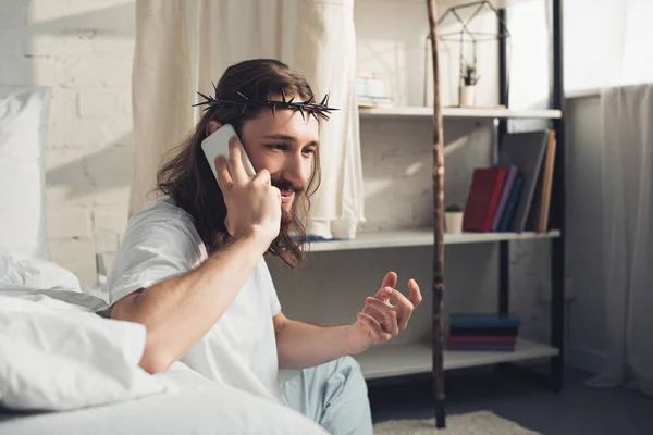 Веселый Иисус в терновом венце разговаривает по смартфону в спальне дома — стоковое фото