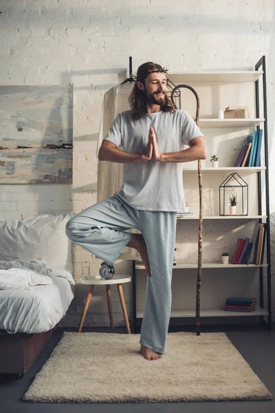 Smiling Jesus in crown of thorns standing in tree pose in bedroom at home — Stock Photo