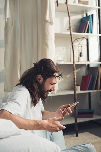 Боковой вид Иисуса в терновом венце с помощью смартфона в спальне дома — стоковое фото