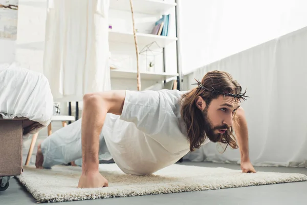 Selective focus of Jesus in crown of thorns doing push ups during morning time in bedroom at home — Stock Photo