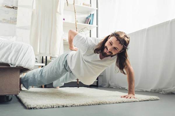 Smiling Jesus in crown of thorns doing push ups on one hand during morning time in bedroom at home — Stock Photo