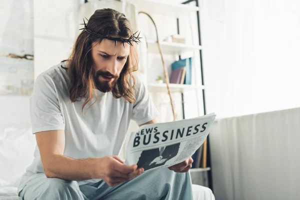 Serious Jesus in crown of thorns reading business newspaper in bedroom during morning time at home — Stock Photo
