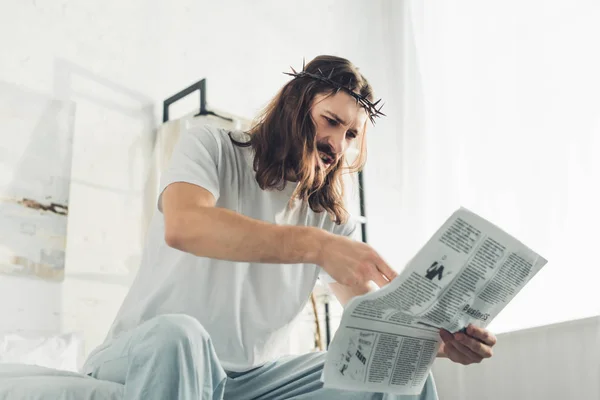 Jesus irritado na coroa de espinhos lendo jornal de negócios no quarto durante o horário da manhã em casa — Fotografia de Stock