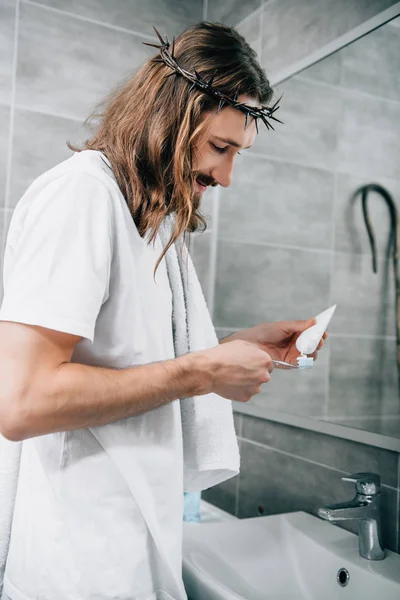 Side view of Jesus in crown of thorns putting toothpaste on brush in bathroom — Stock Photo