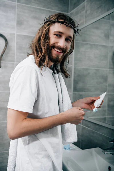 Glücklicher Jesus in Dornenkrone blickt in die Kamera und legt Zahnpasta auf Bürste im Badezimmer — Stock Photo