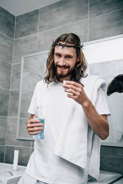 Jesús alegre en corona de espinas sosteniendo el enjuague bucal y mirando a la cámara en el baño - foto de stock