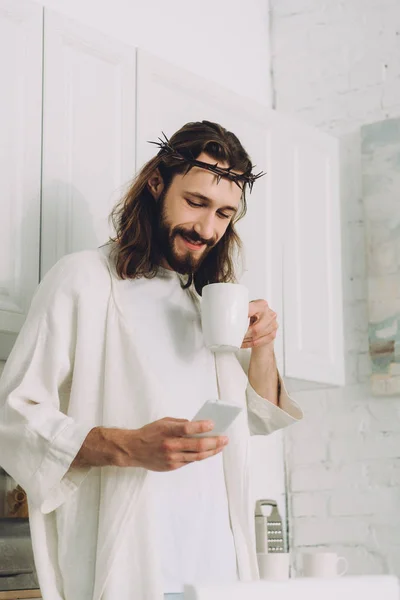 Enfoque selectivo de Jesús en la corona de espinas de pie con taza de café y el uso de teléfono inteligente en la cocina en casa - foto de stock