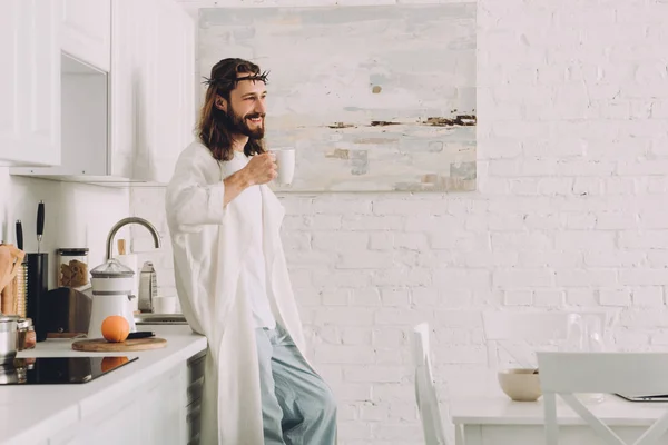 Cheerful Jesus in crown of thorns standing with cup of coffee in kitchen at home — Stock Photo