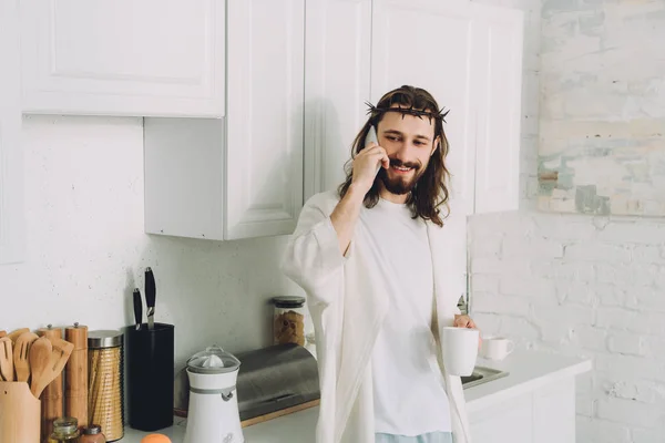 Smiling Jesus in crown of thorns standing with cup of coffee and talking on smartphone in kitchen at home — Stock Photo