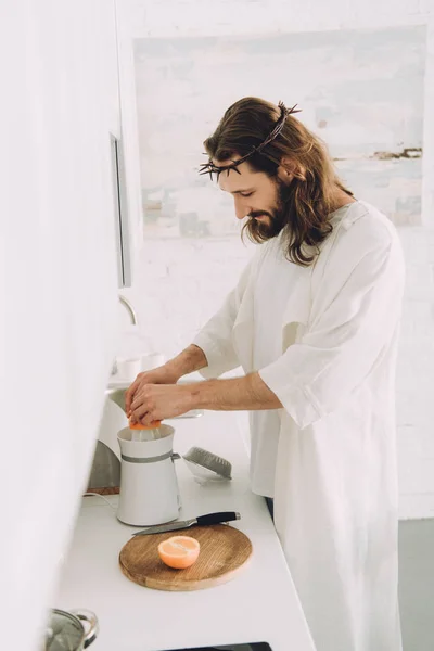 Side view of cheerful Jesus doing orange juice by juicer in kitchen at home — Stock Photo
