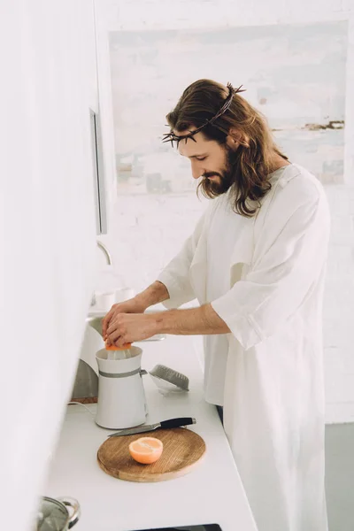 Vista lateral de Jesús alegre haciendo jugo de naranja por exprimidor en la cocina en casa — Stock Photo