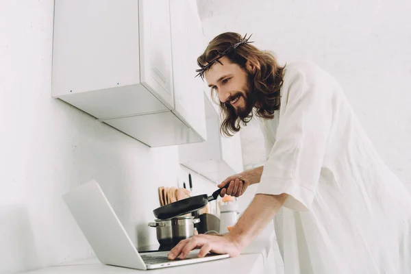 Sonriendo a Jesús usando el ordenador portátil mientras se cocina con sartén en la cocina en casa - foto de stock