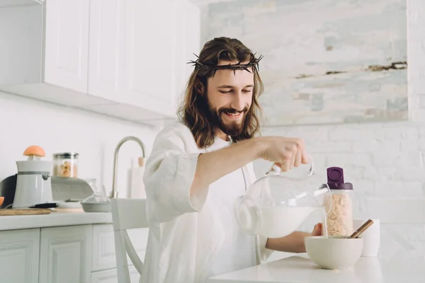 Feliz Jesús vertiendo leche en un tazón con hojuelas de maíz en el desayuno en la cocina en casa - foto de stock