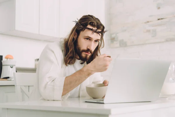 Focado Jesus comer flocos de milho no café da manhã à mesa com laptop na cozinha em casa — Fotografia de Stock