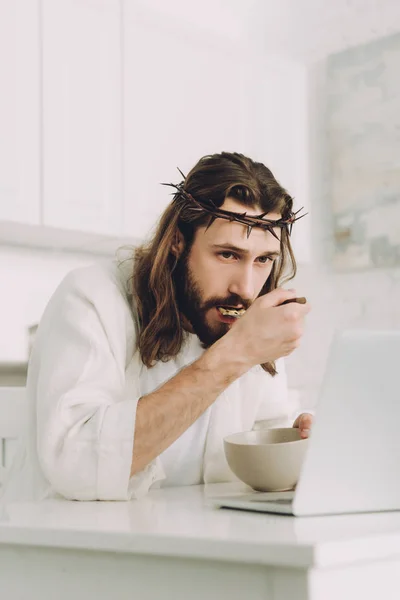 Selective focus of Jesus eating corn flakes on breakfast at table with laptop in kitchen at home — Stock Photo