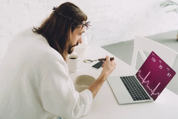 Concentrated Jesus eating corn flakes on breakfast at table with laptop with graph on screen in kitchen at home — Stock Photo