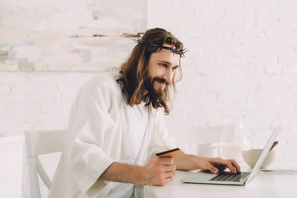 Alegre Jesús sosteniendo la tarjeta de crédito y haciendo compras en línea en la mesa con el ordenador portátil en la cocina en casa - foto de stock