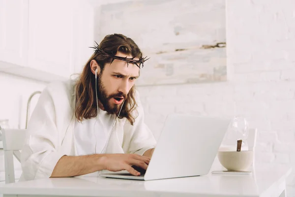 Emotional Jesus in crown of thorns and earphones using laptop at table in kitchen at home — Stock Photo