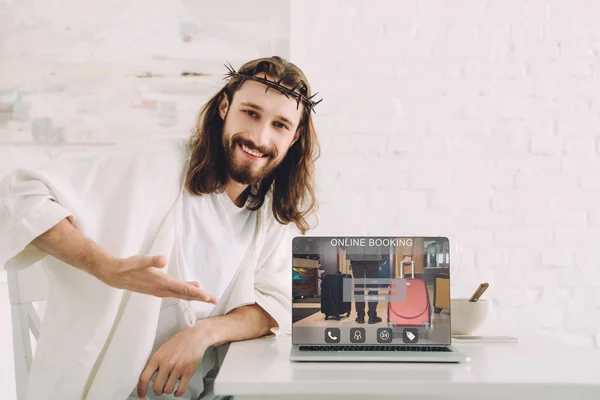 Smiling Jesus in crown of thorns pointing at laptop with online booking website in kitchen at home — Stock Photo