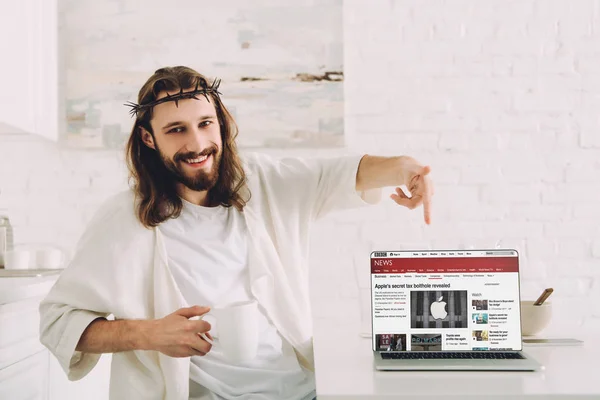 Cheerful Jesus holding cup of coffee and pointing at laptop with bbc news website in kitchen at home — Stock Photo