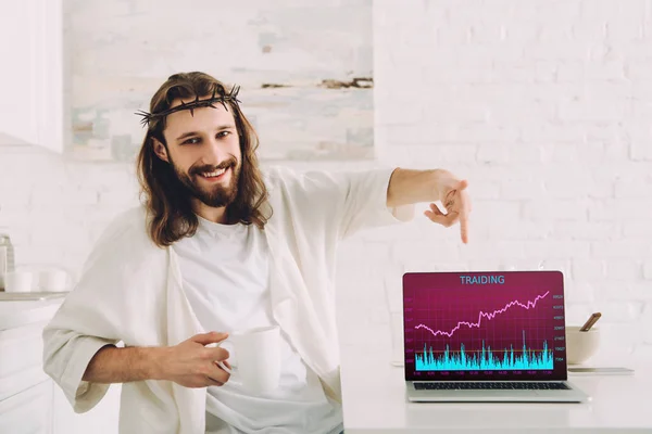 Lächelnder Jesus, der eine Tasse Kaffee in der Hand hält und auf den Laptop zeigt, mit dem Handel auf dem Bildschirm in der heimischen Küche — Stockfoto