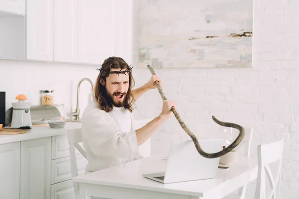 Angry Jesus hitting laptop by wooden staff in kitchen at home — Stock Photo