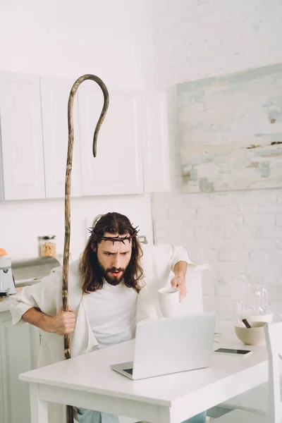 Шокированный Иисус с чашкой кофе и деревянный персонал, глядя на ноутбук на кухне дома — стоковое фото