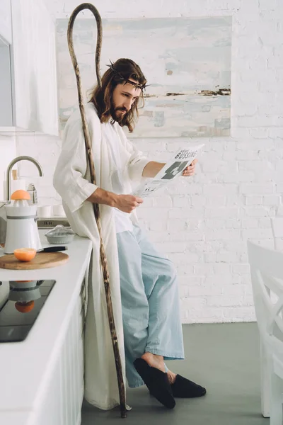 Focused Jesus with wooden staff reading business newspaper in kitchen at home — Stock Photo
