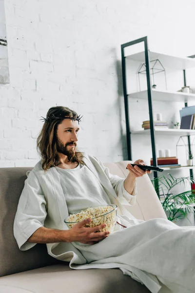 Jesús serio en corona de espinas viendo la televisión y sentado con tazón de palomitas de maíz en el sofá en casa - foto de stock