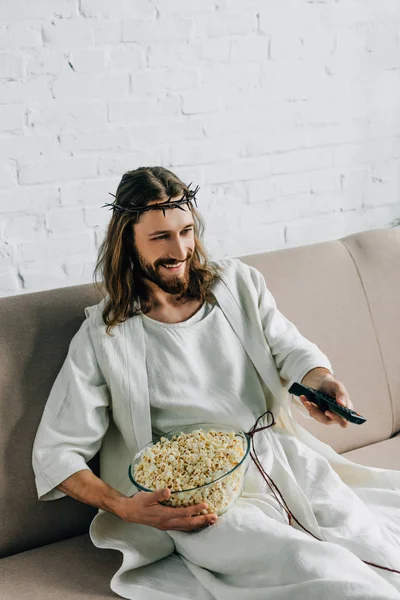 Vue grand angle de Jésus heureux en couronne d'épines regarder la télévision et assis avec bol de maïs soufflé sur le canapé à la maison — Photo de stock