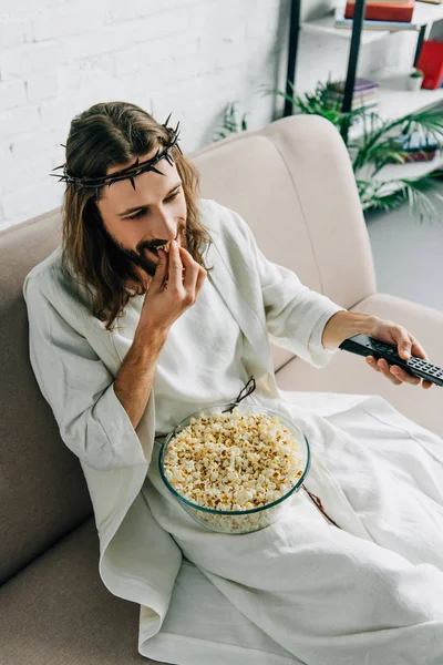 Vue grand angle de Jésus en couronne d'épines regarder la télévision et manger du pop-corn sur le canapé à la maison — Photo de stock