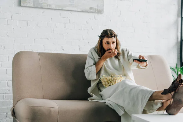 Selective focus of Jesus in crown of thorns watching tv and eating popcorn on sofa at home — Stock Photo