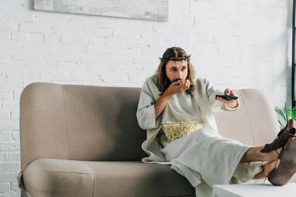 Foco seletivo de Jesus na coroa de espinhos assistindo tv e comer pipocas no sofá em casa — Fotografia de Stock