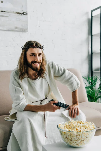 Sorrindo Jesus na coroa de espinhos assistindo tv e sentado com tigela de pipoca no sofá em casa — Fotografia de Stock