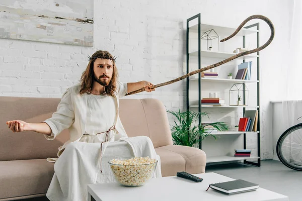 Serious Jesus sitting on sofa with wooden staff and watching tv at home — Stock Photo
