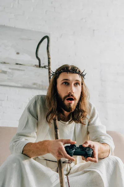 Emotional Jesus with wooden staff playing video game by joystick on sofa at home — Stock Photo