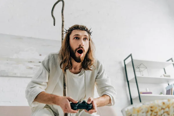 Шокированный Иисус с деревянным посохом, играющим в видеоигры на диване дома — стоковое фото