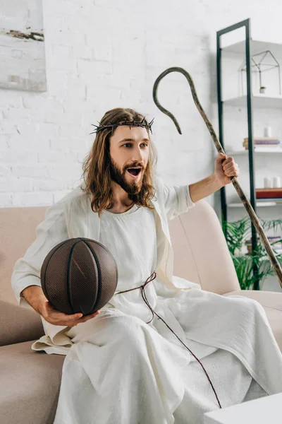 Cheerful Jesus with wooden staff holding ball and watching basketball match on sofa at home — Stock Photo